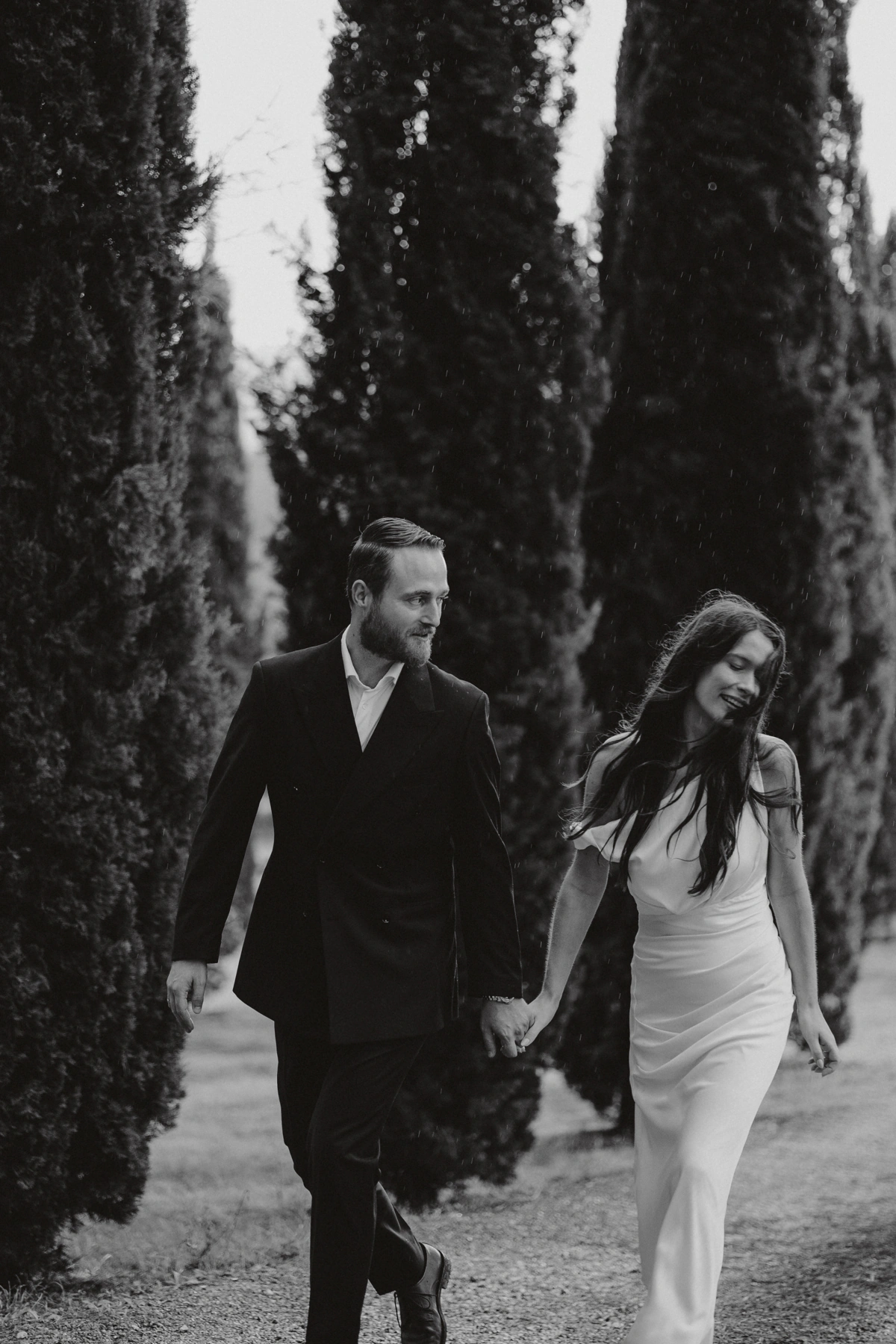 Sposi che camminano mano nella mano tra i cipressi, in bianco e nero — fotografia di Andrea Mortini